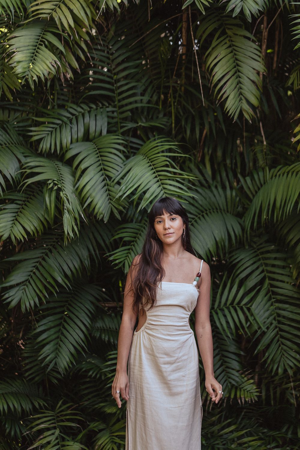 A woman in a cream colored dress poses against a backdrop of lush tropical palm fronds in a garden setting.