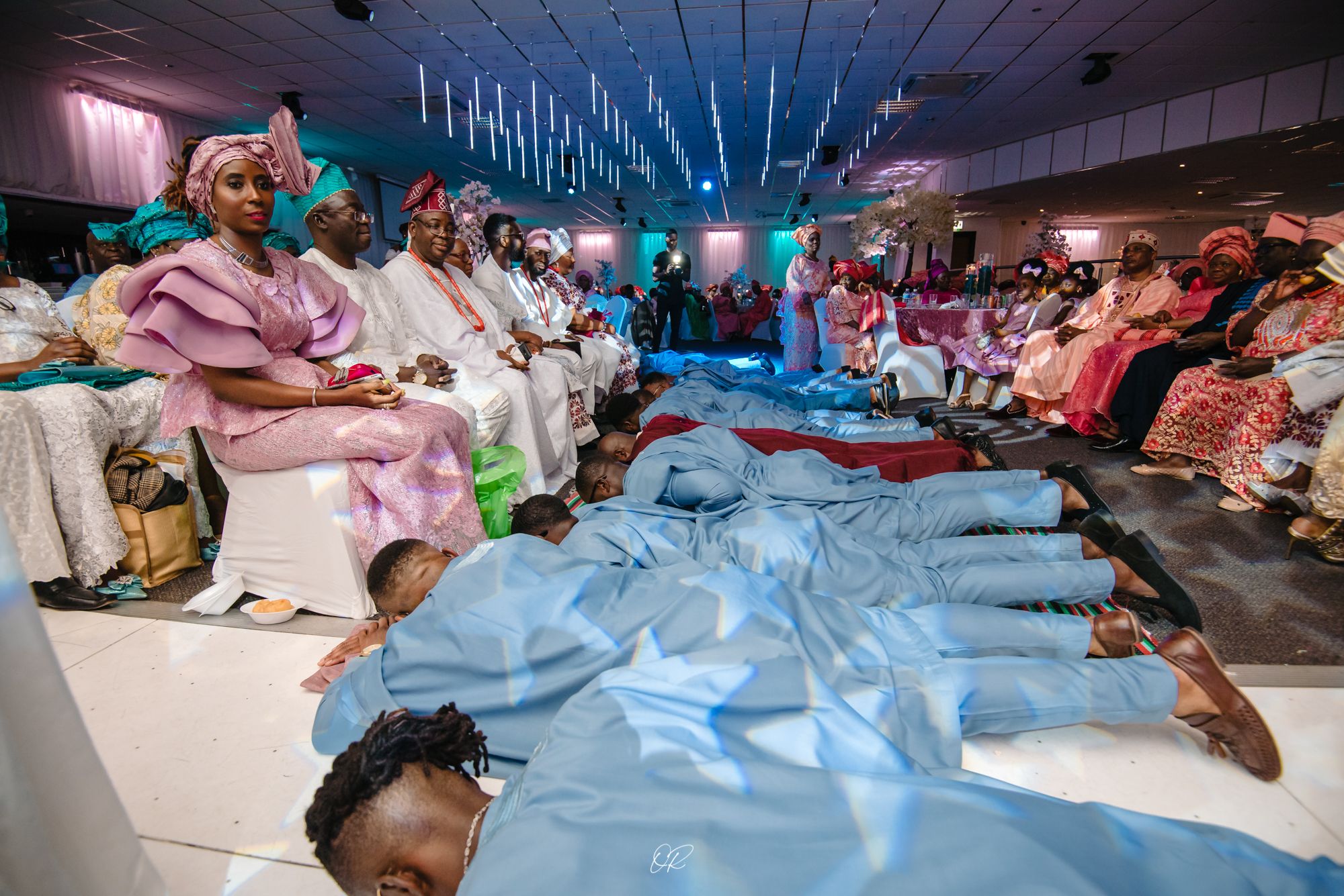 Wedding guests in traditional attire sitting in a modern venue with blue lighting and flowing fabric decor.