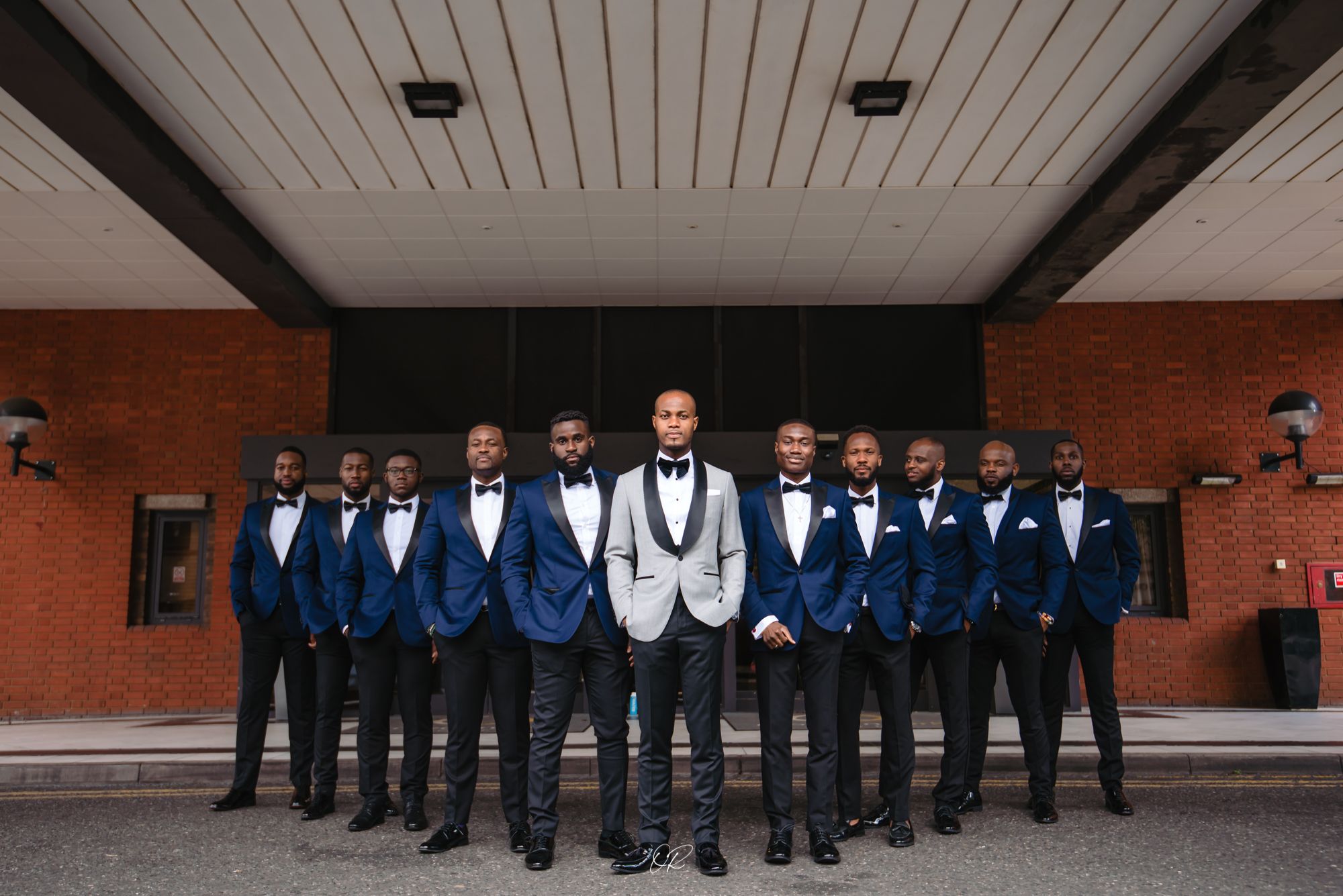 Wedding party in matching blue suits and black pants pose together on brick building entrance steps.