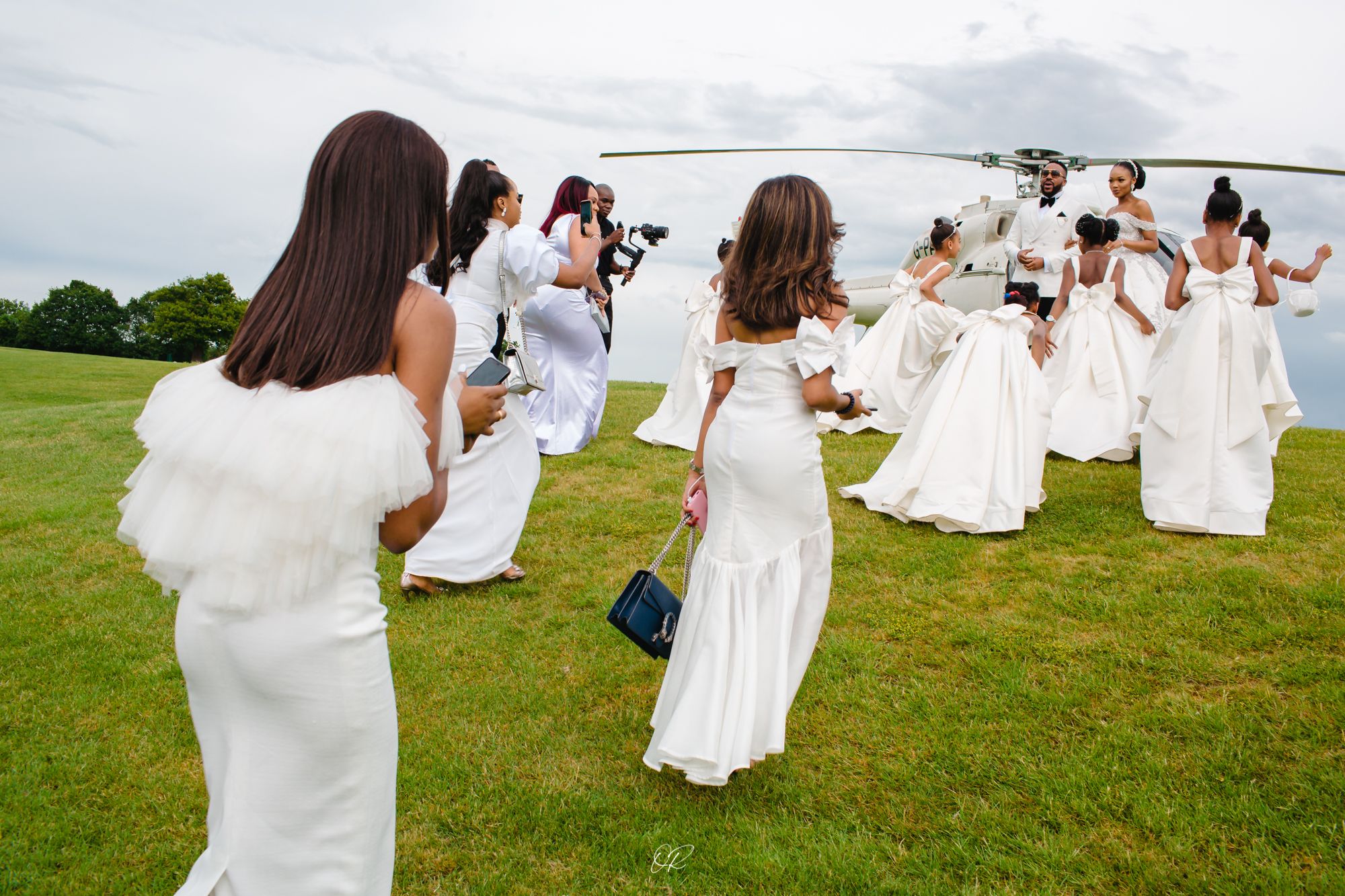 Photographers capture a group of people in white dresses posing on a grassy hilltop.