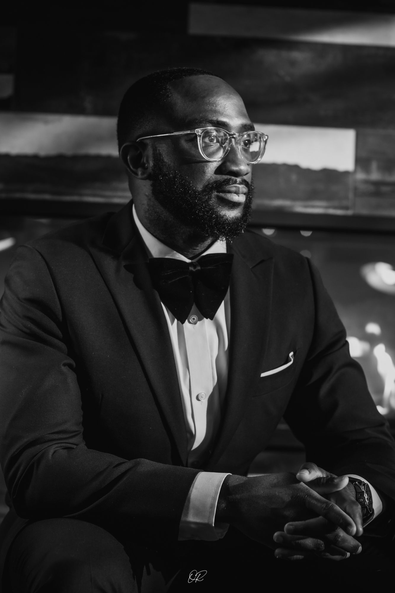Black and white portrait of a person in a formal tuxedo with bow tie and glasses looking contemplative.