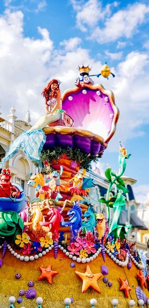 Colorful mermaids and sea creatures decorate a whimsical carnival float with pearls and shells against a blue sky.