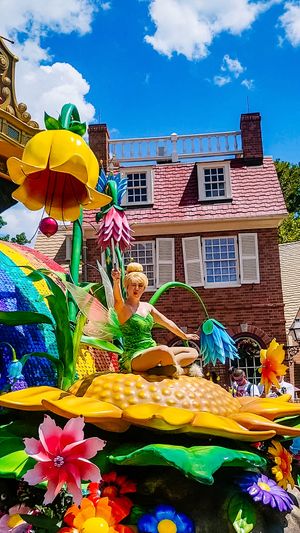 Colorful parade float with giant flowers and fantasy elements passing by a colonial-style house under bright blue sky.