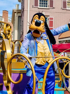 Pluto character wearing blue overalls, yellow vest and denim jacket on a colorful parade float at Disney theme park.