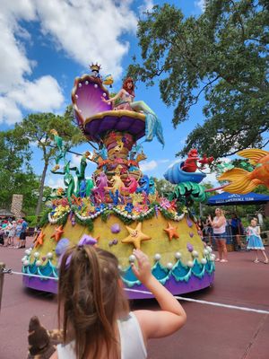 A colorful Disney parade float with ocean and mermaid themes passing through Magic Kingdom as guests watch the spectacle.