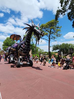 Panoramic view of a Disney park pathway in Magic Kingdom with park guests and a large metal dragon sculpture nearby.