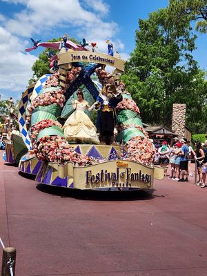 A colorful Festival of Fantasy parade float with flowers and decorative elements moves through a Disney theme park on a sunny day.