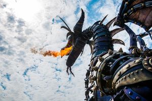 A large robotic dragon sculpture breathing flames against a cloudy sky during an outdoor display performance.