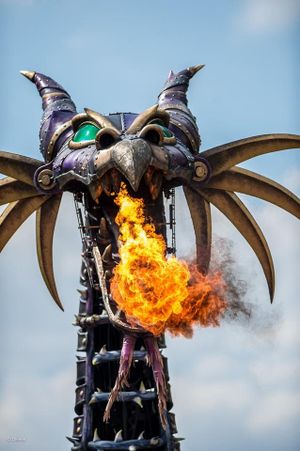 A robotic dragon sculpture mounted on a pole spits out flames against a blue sky with dramatic smoke effects.