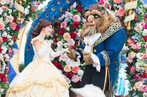 Two characters in ornate costumes reminiscent of Beauty and the Beast pose together against a floral archway backdrop.