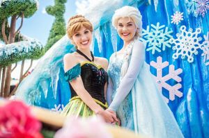 Costumed performers in princess dresses pose together in front of bright blue winter-themed backdrops at a theme park.