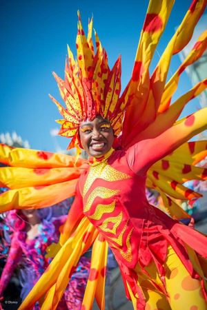 Vibrant carnival performer in red and yellow sunburst costume with dramatic feathered headdress dances against blue sky.