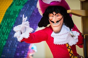 An animated mascot character in a red coat, white ruffles and pink feathered hat poses energetically against a rainbow backdrop.