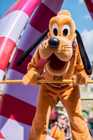 Pluto the Disney character performs in a street parade at a theme park, holding drumsticks against a red and white backdrop.