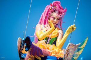 Pink-haired performer dances with yellow fabric streamers against a vibrant blue background.