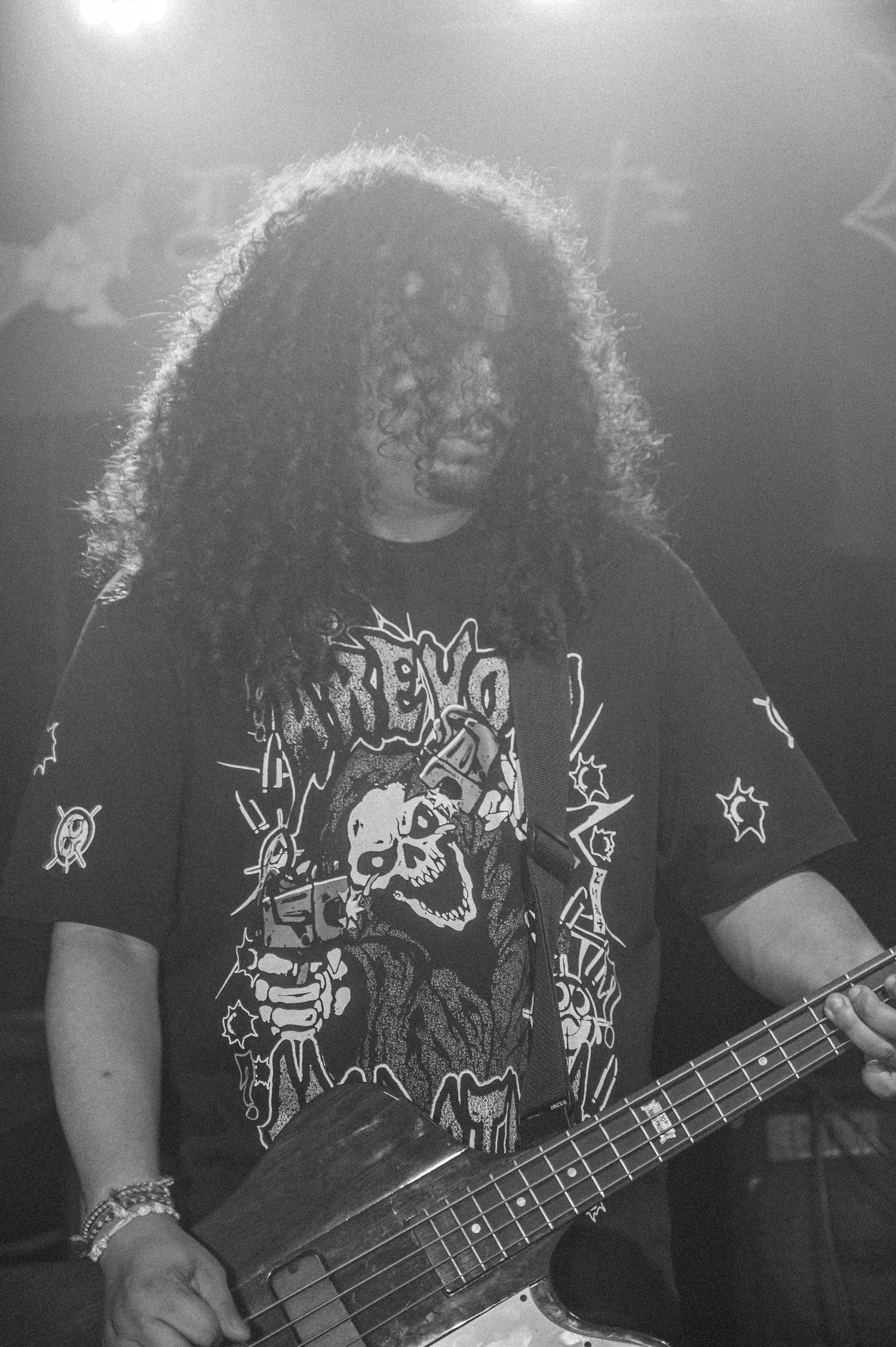 Black and white photo of a bass player performing on stage wearing a metal band t-shirt with skull artwork.