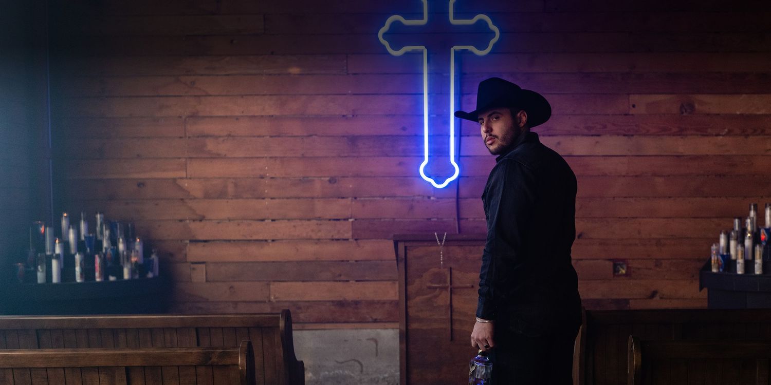 A silhouetted figure in a black hat stands against a wooden wall with a glowing blue neon cross and lit candles.
