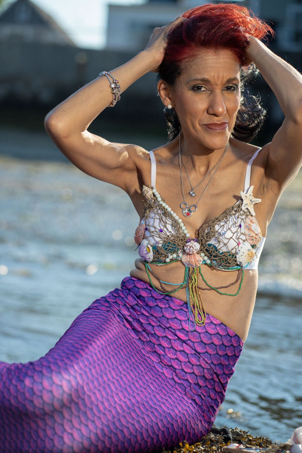 A person with red hair poses in a purple mermaid tail costume and shell top by the water.