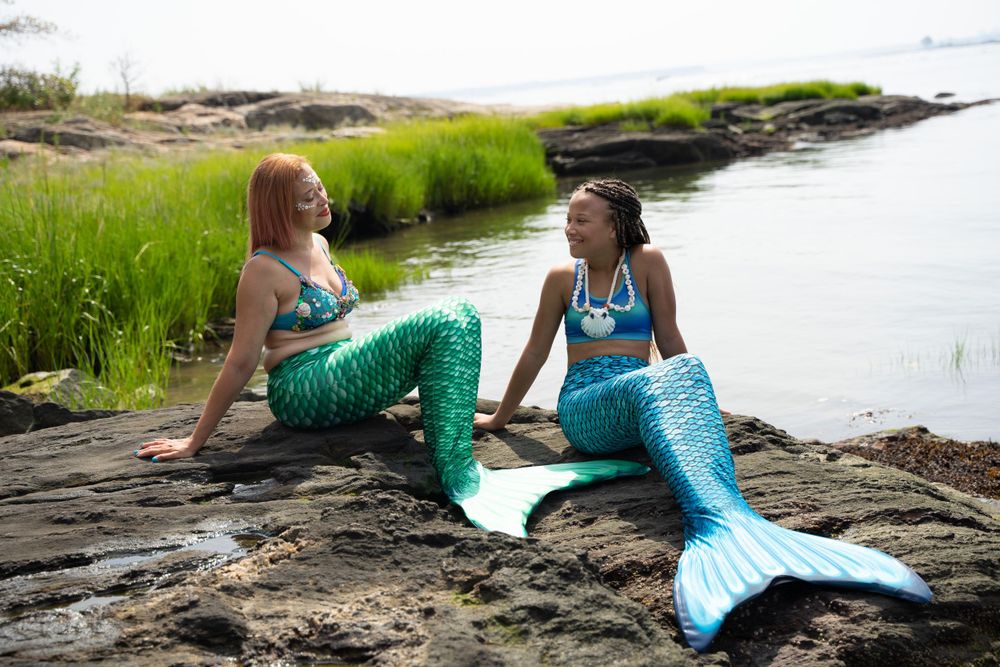 Two people in turquoise mermaid costumes lounging on coastal rocks near marsh grass.