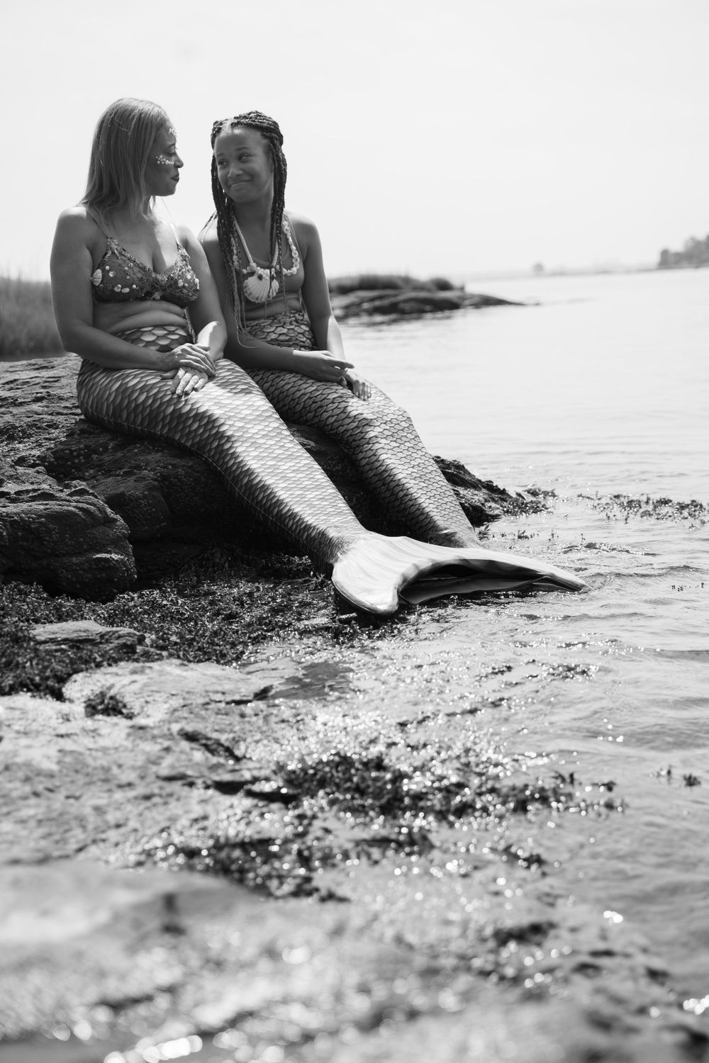 Black and white photo of two people in mermaid costumes relaxing by the water.