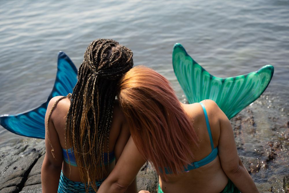 Two people wearing mermaid tail costumes sitting together on rocks by the ocean.