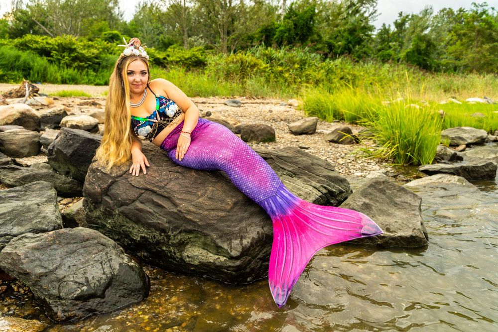 Model in floral top and pink mermaid tail costume reclining on coastal rocks near water.