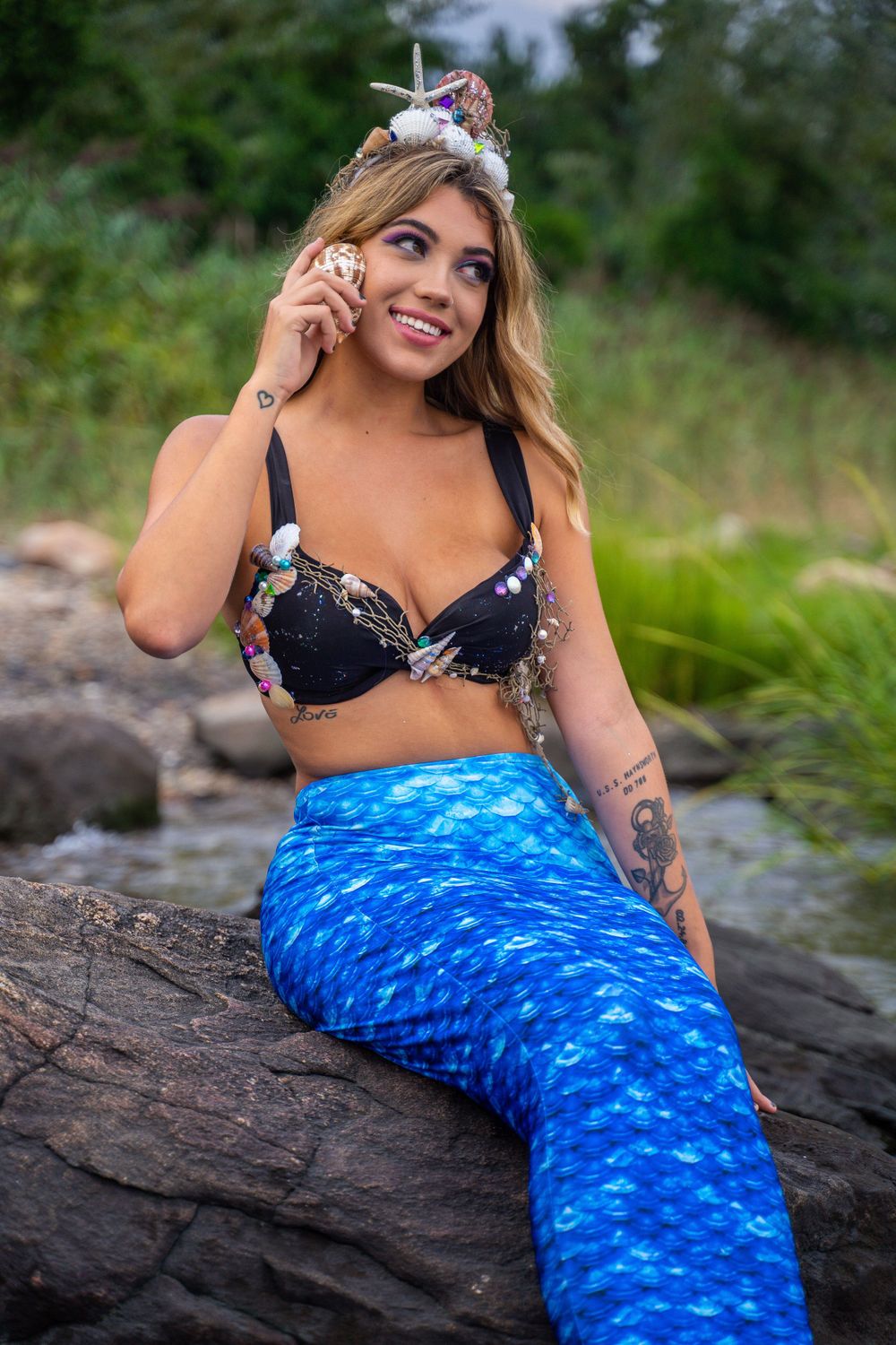Person in a floral bikini top with mermaid tail costume posing on rocky shore by water.