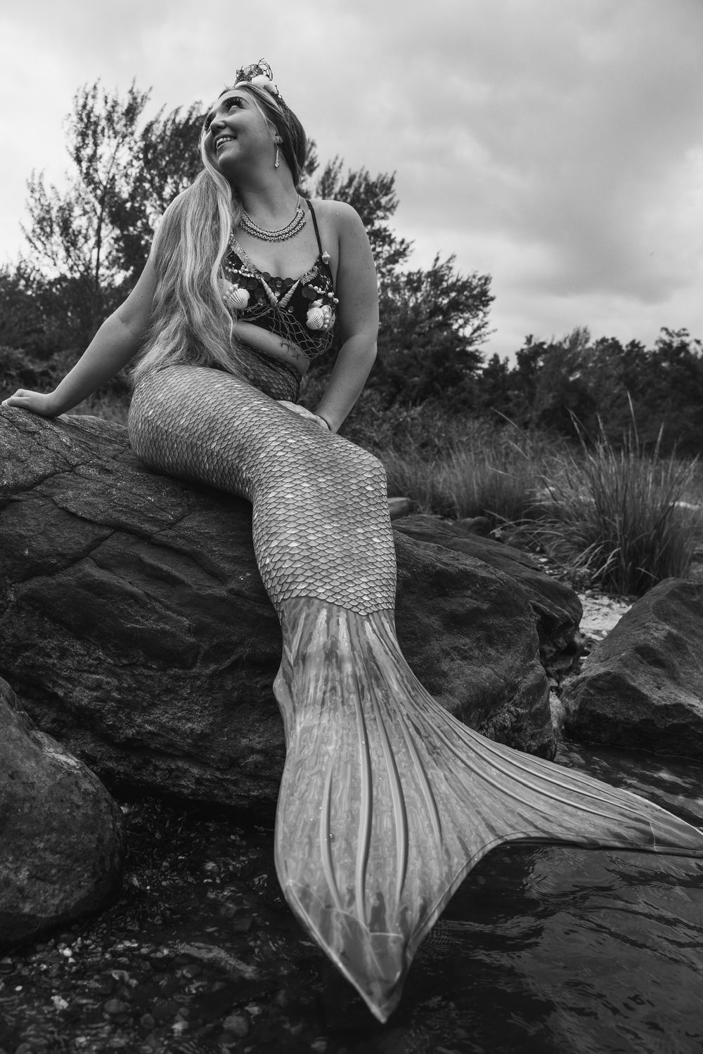 Black and white portrait of person in sparkly mermaid tail costume sitting on coastal rocks.