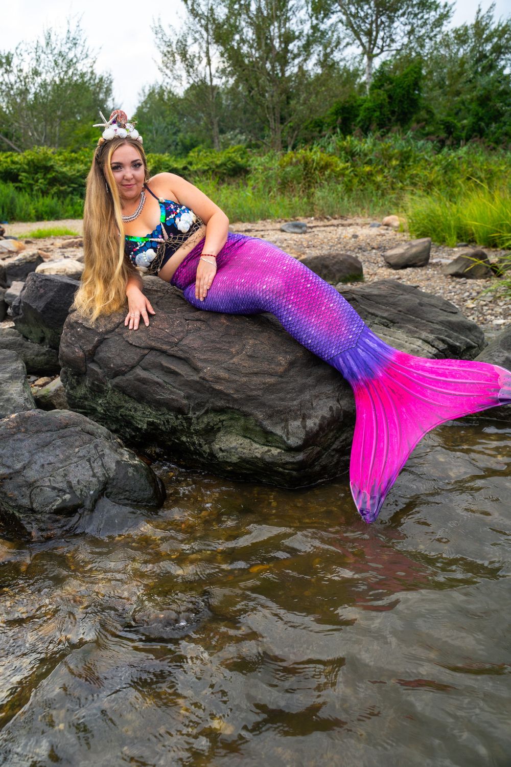 Model wearing purple and pink gradient mermaid tail costume poses on rocky shoreline.