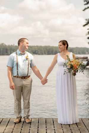 A couple in wedding attire holds hands on a wooden dock by a serene lake.