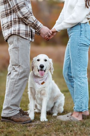 White fluffy dog sitting between couple's legs in autumn field.