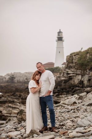 A couple shares an intimate moment in front of a lighthouse on a rocky coastal shoreline.