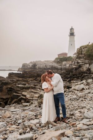 A tender moment between a couple on rocky shores with a white lighthouse towering above.