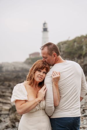 An intimate portrait of a couple embracing near a lighthouse on a foggy coastline.
