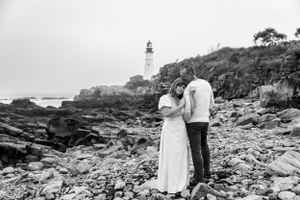 A black and white photograph of a couple sharing a moment by a coastal lighthouse.