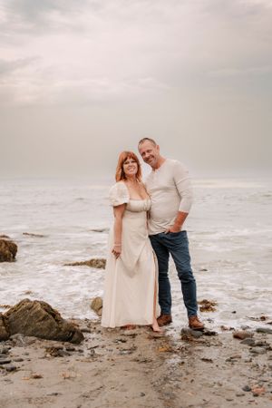 A couple stands together on a beach with crashing waves and cloudy skies in the background.