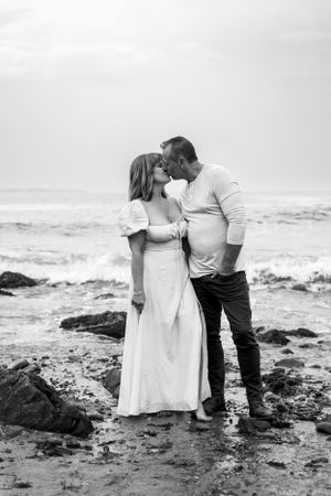A black and white beach portrait of a couple sharing an intimate moment among coastal rocks.