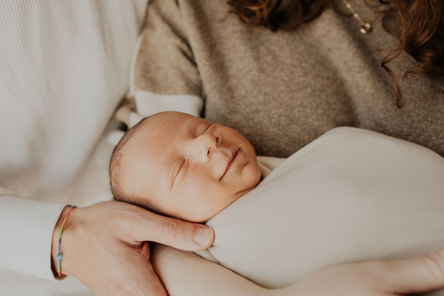 A sleeping newborn baby rests peacefully in gentle arms against a soft beige background.