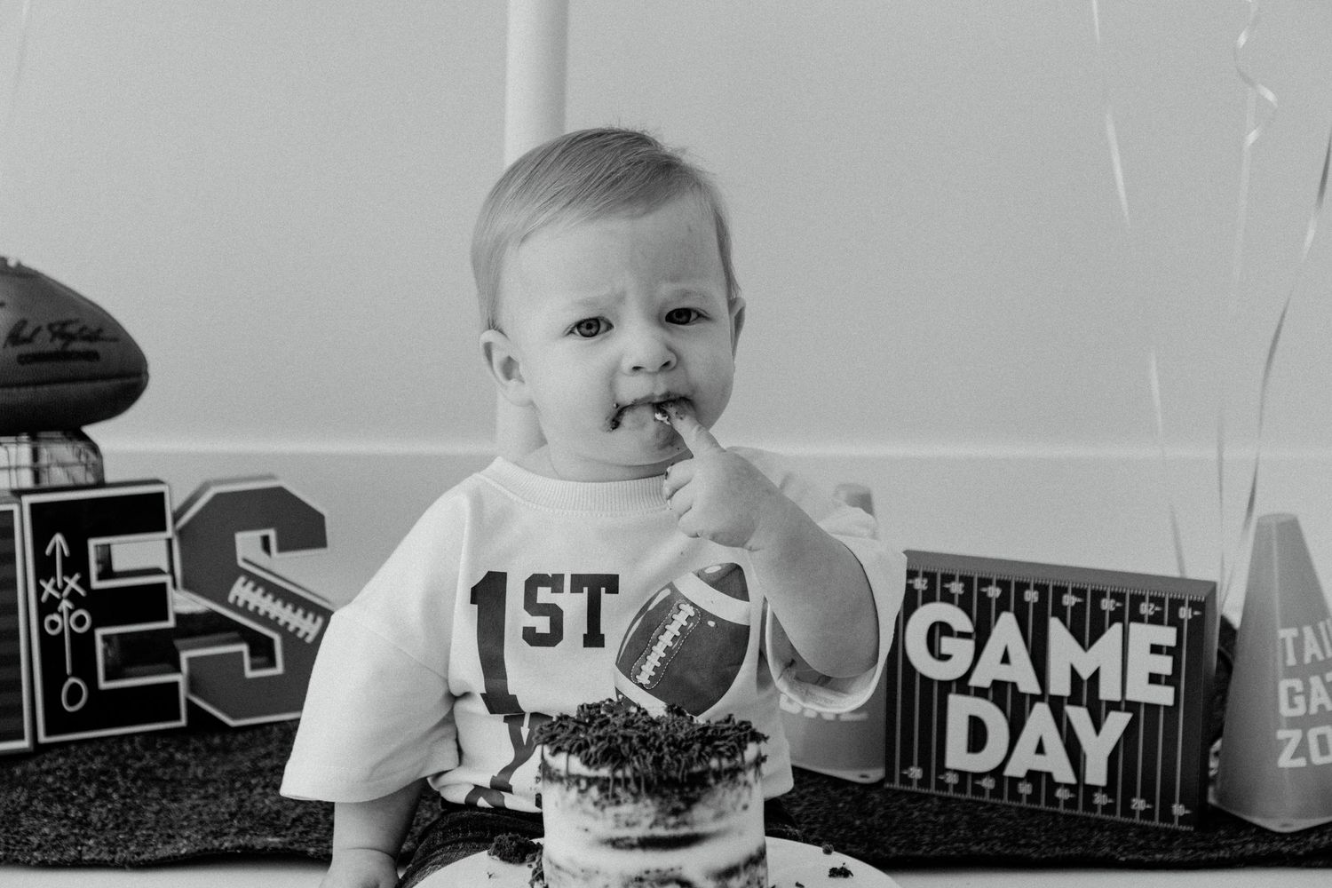 A baby in a white shirt holds a football during their first game day celebration in black and white.