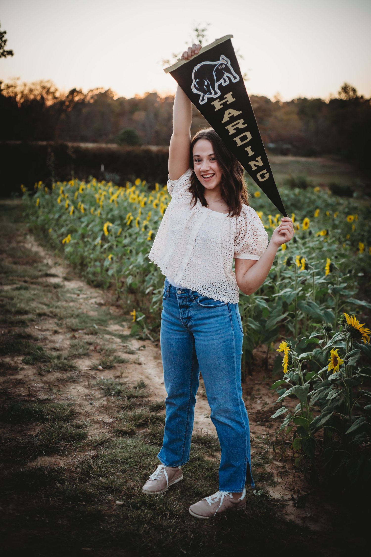 Graduate posing with celebratory pennant flag in casual attire against a beautiful sunflower field at sunset.