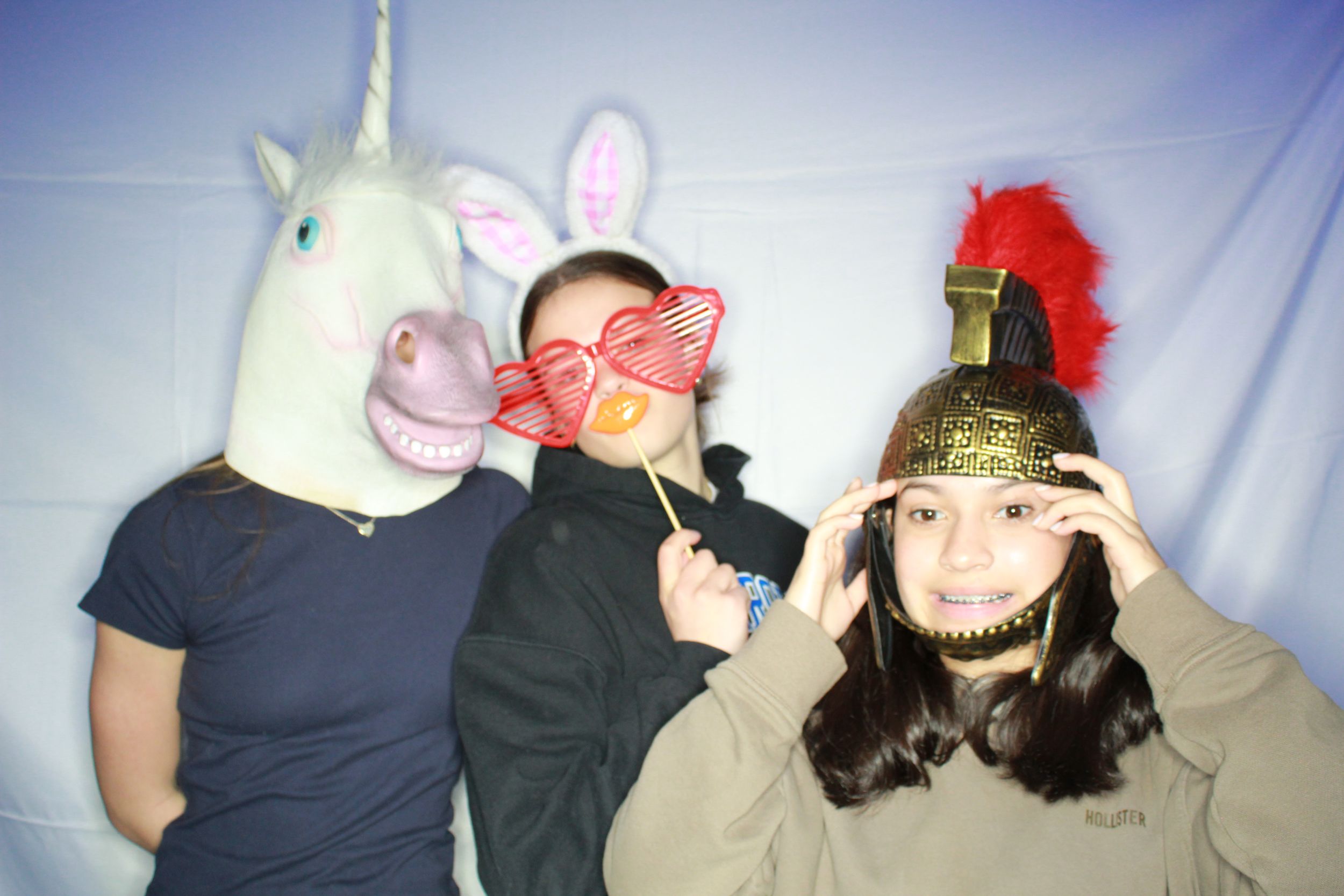 Three people pose for a fun photo booth shot wearing costumes including a unicorn mask, red heart glasses and a festive hat.