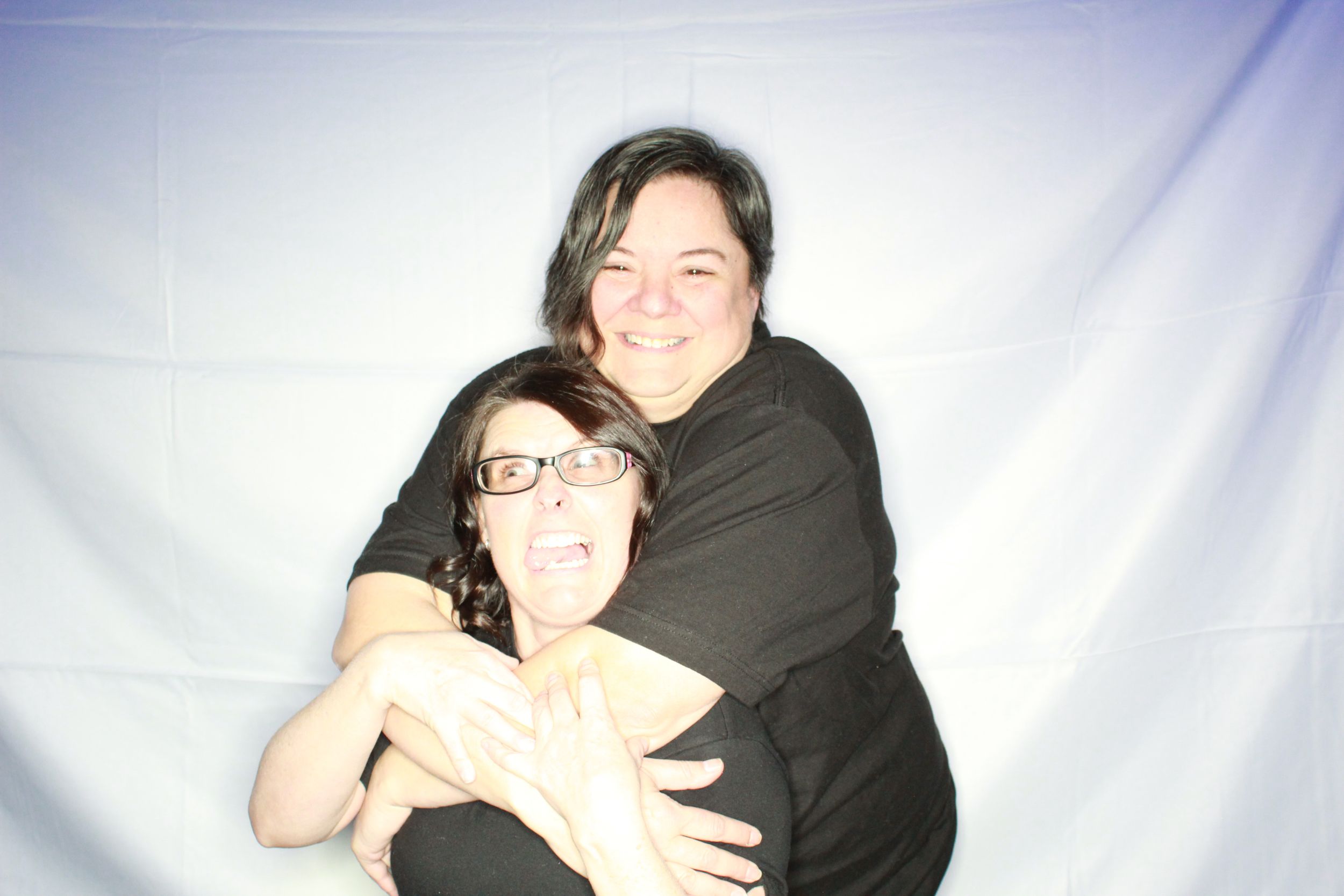 Two people in black clothing embracing and smiling against a light colored background in a photo booth style shot.