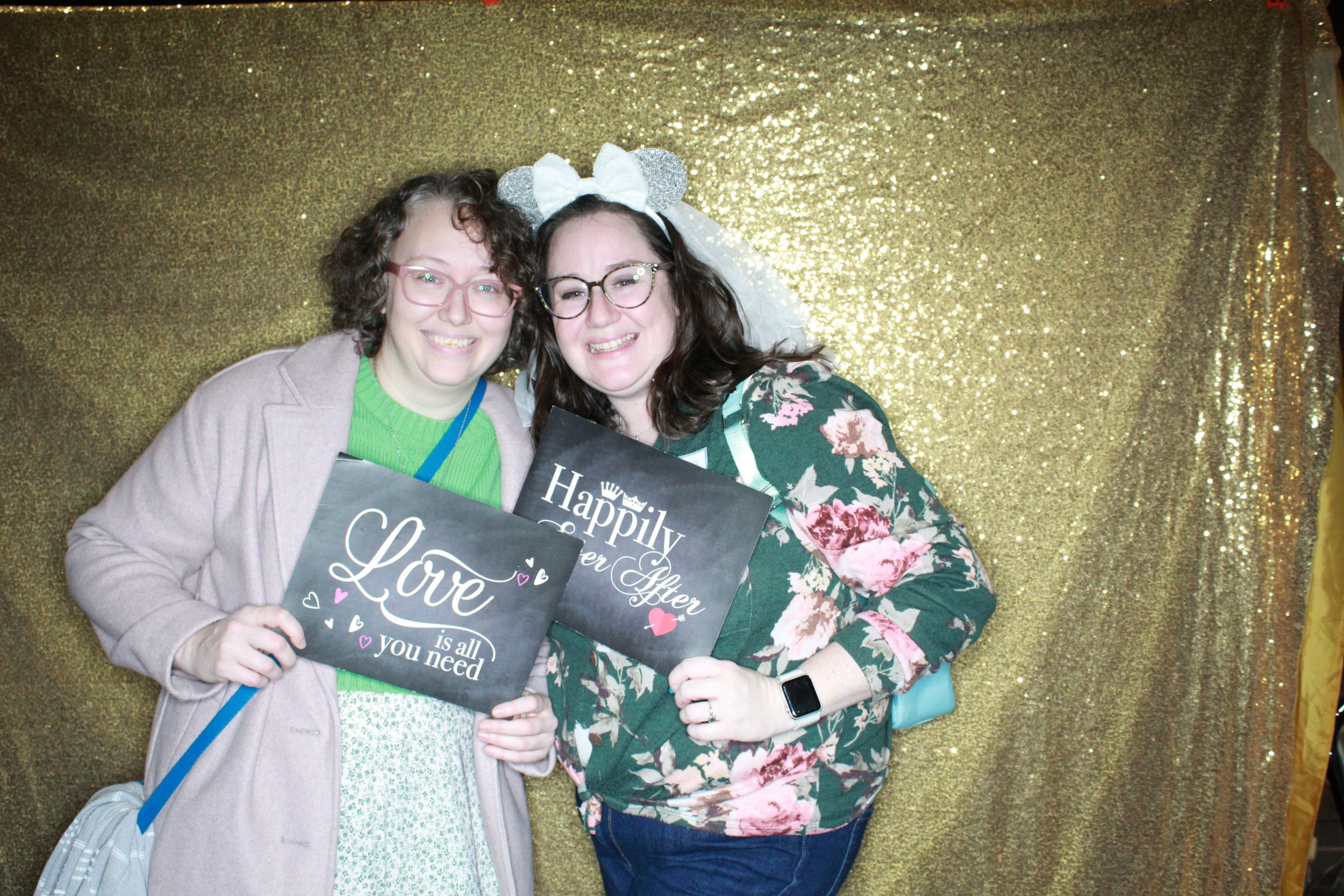 Two people posing with a sparkly gold backdrop while holding a black chalkboard sign at a holiday event.