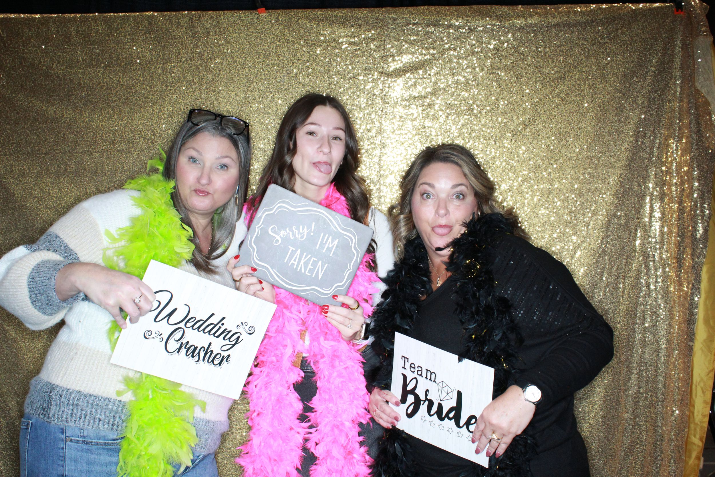 Three people pose with wedding-themed signs and colorful feather boas against a gold backdrop at a photo booth party.