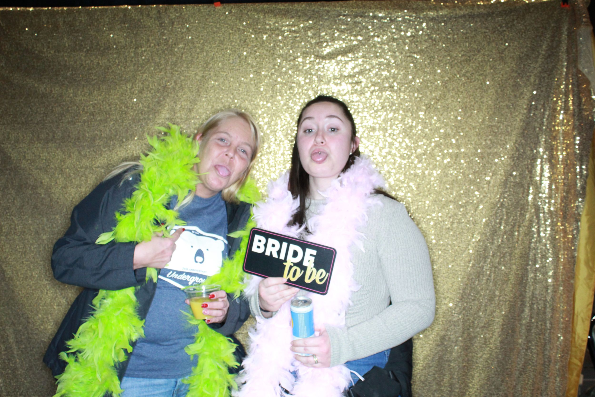 Two people pose with bridal signs and a bright green feather boa against a gold shimmer backdrop at a party.