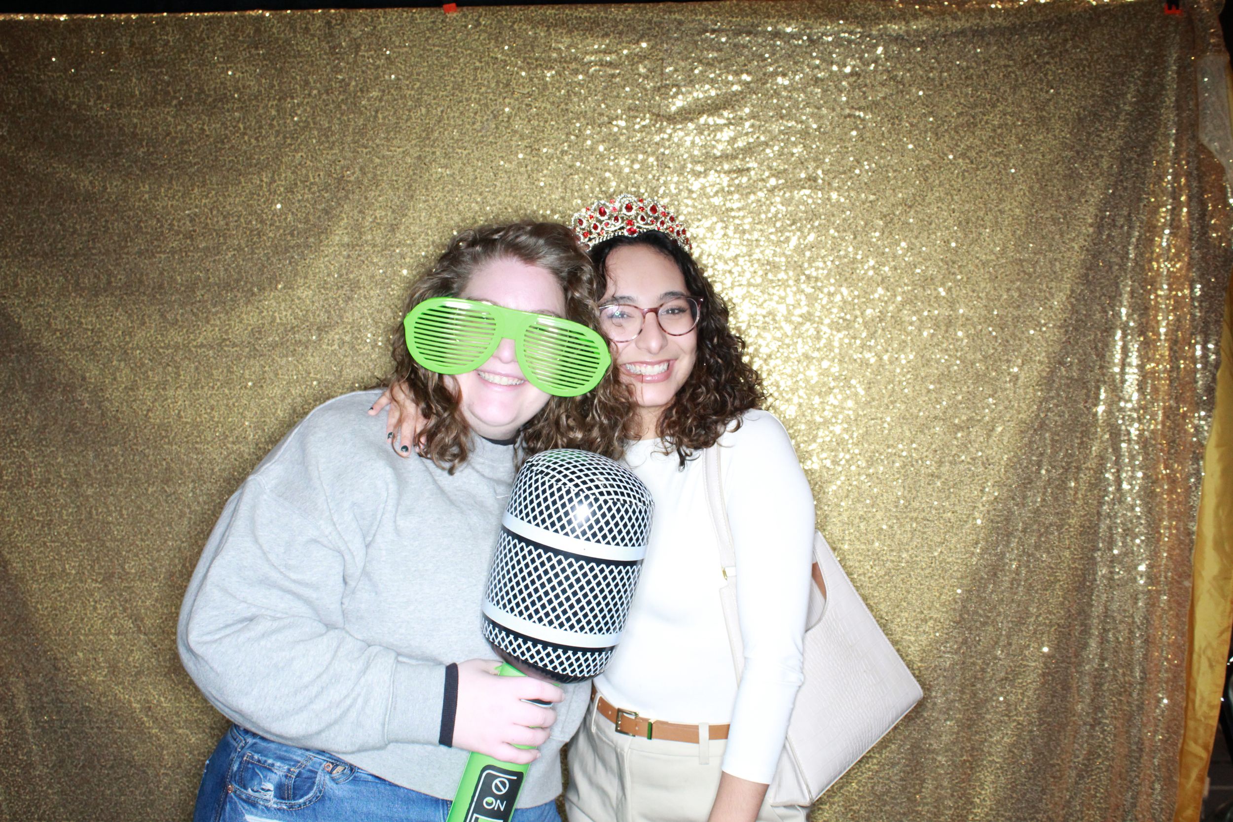 Two people pose together against a gold backdrop with fun party props including an inflatable microphone and green glasses.