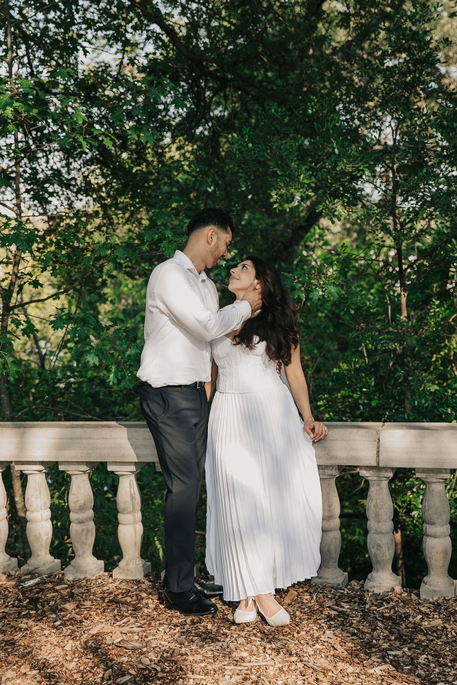 A romantic couple embraces outdoors, smiling at each other amid sunlit foliage in the background.