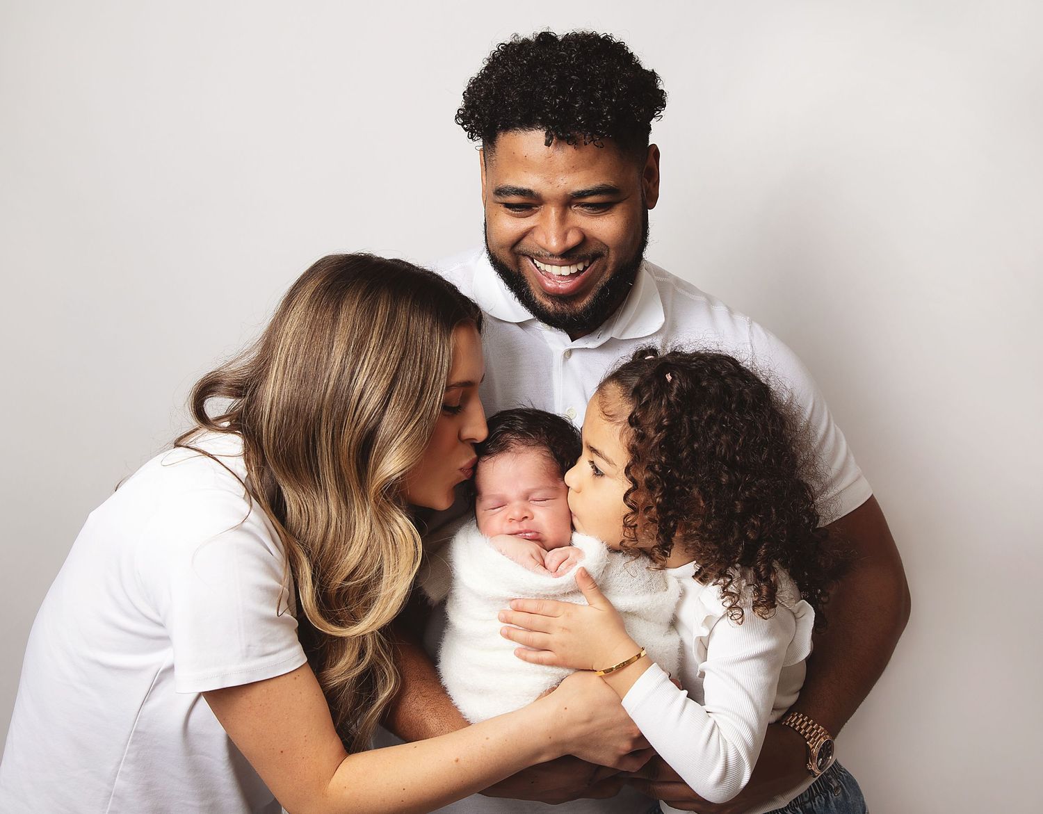 A loving family in white shirts sharing an intimate moment with their newborn baby against a light background.