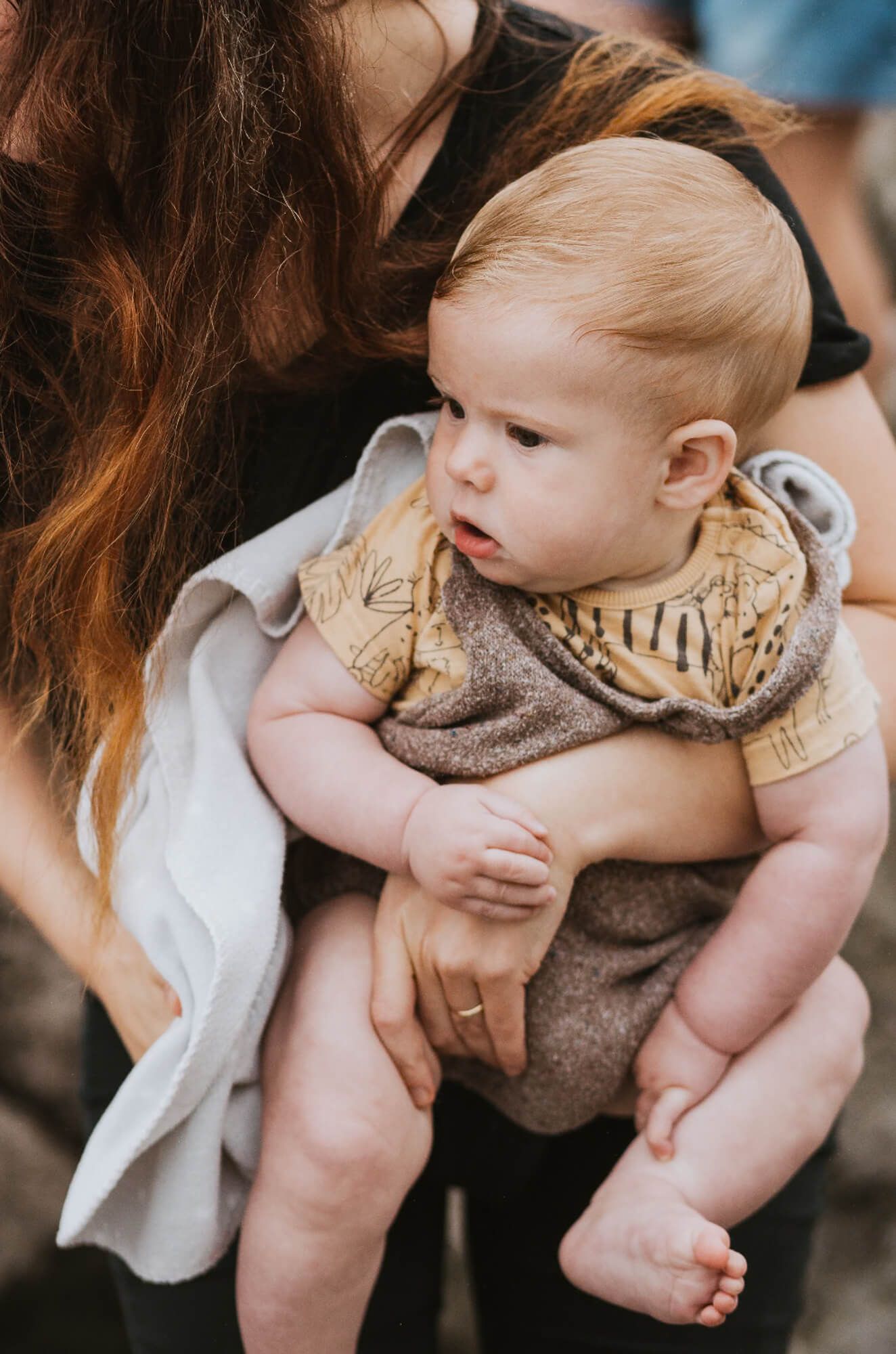 Baby wearing beige romper sits in caregiver's lap while looking to the side with curious expression.