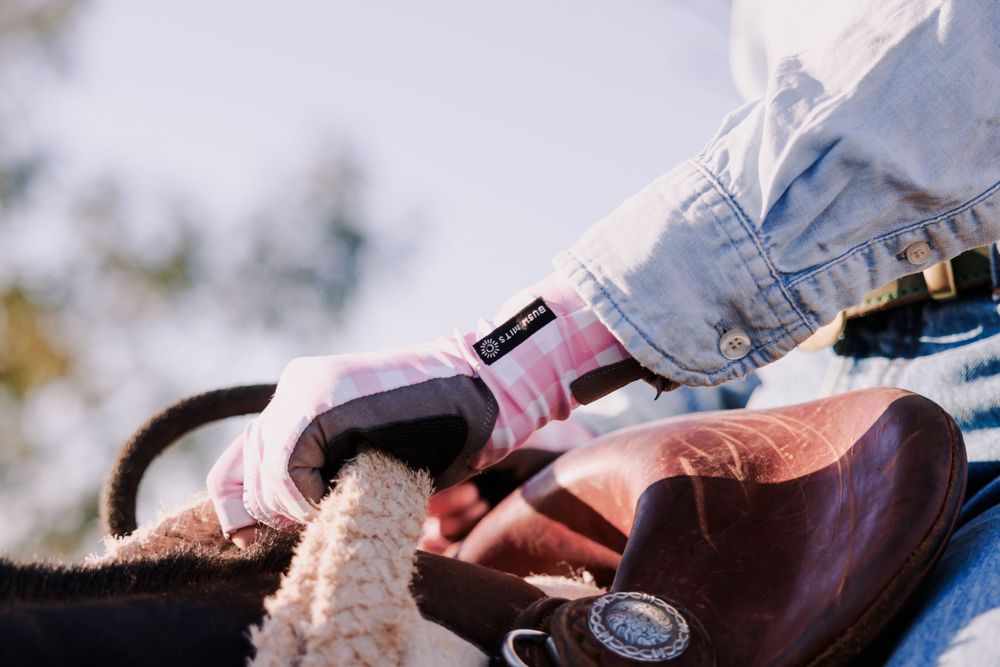 Close-up of hands wearing pink gloves adjusting a brown leather saddle with sheepskin lining.